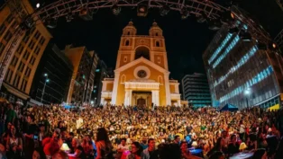 Público reunido em praça durante apresentação noturna em frente a uma igreja, cercada por prédios.