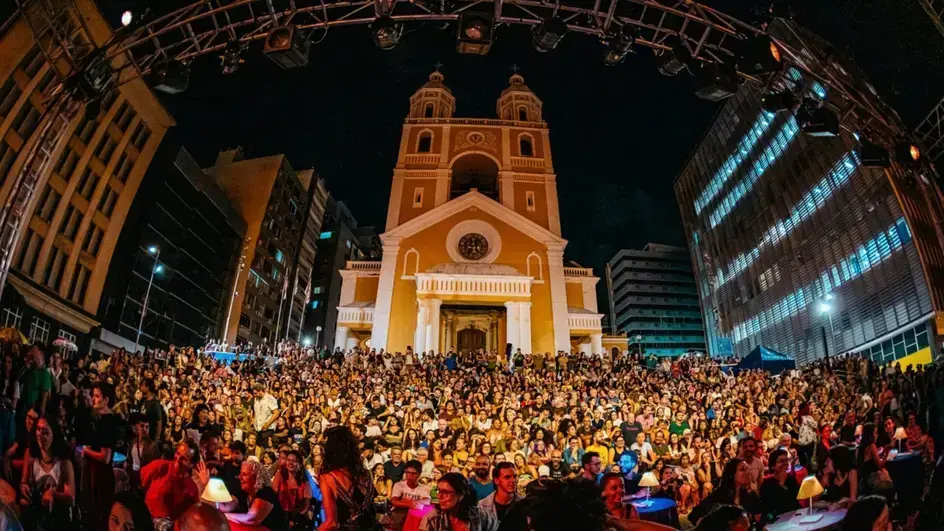 Público reunido em praça durante apresentação noturna em frente a uma igreja, cercada por prédios.