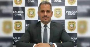 Retrato de um homem com terno, sorrindo e posando em frente a um fundo com logotipos da Polícia Civil.