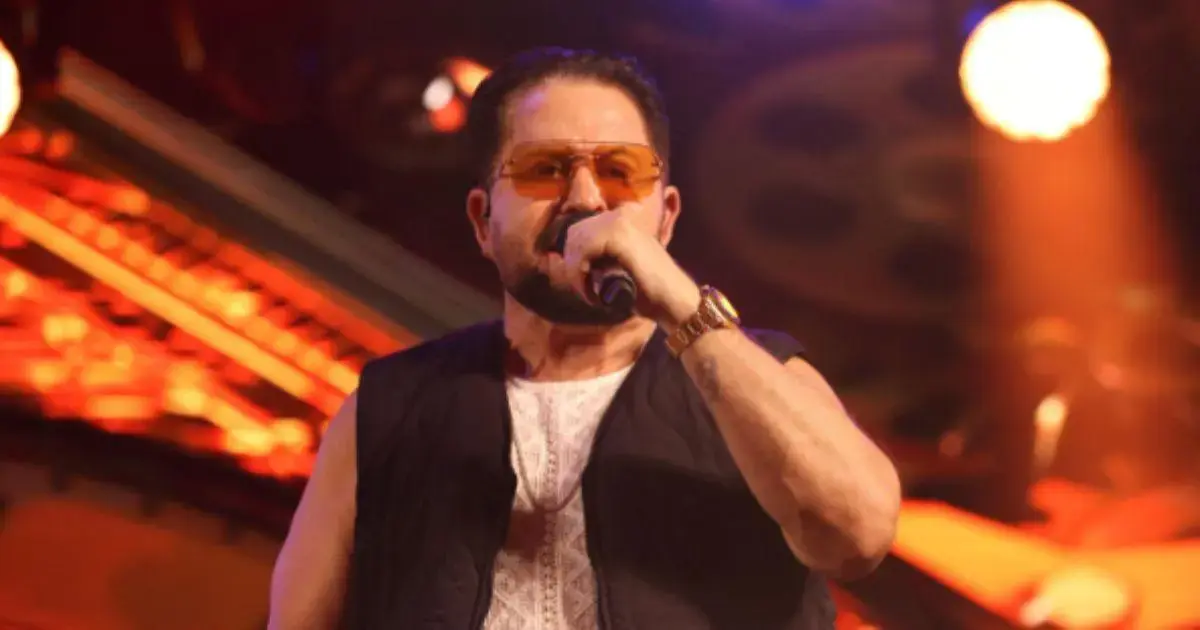 Artista em performance no palco, segurando um microfone e usando óculos escuros, sob luzes coloridas.