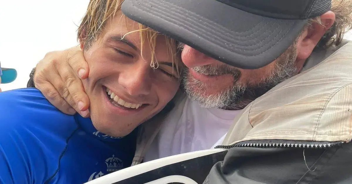 Dois homens se abraçam, sorrindo, em um momento de alegria e celebração, com um surfboard em destaque.