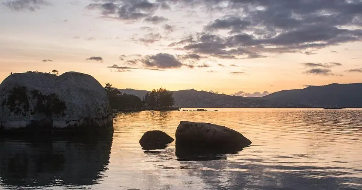 Pôr do sol refletido na água, com grandes rochas à beira de um lago e montanhas ao fundo.