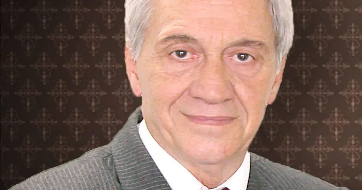 Retrato de um homem de cabelos grisalhos, usando um paletó cinza e com fundo decorativo escuro.