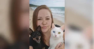 Pessoa segurando um cachorro preto e um gato branco, com a praia ao fundo e céu parcialmente nublado.