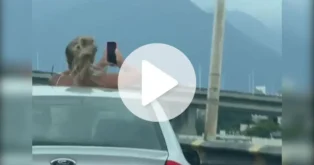 Mulher em pé no teto solar de um carro, segurando um celular, com vista para montanhas ao fundo.