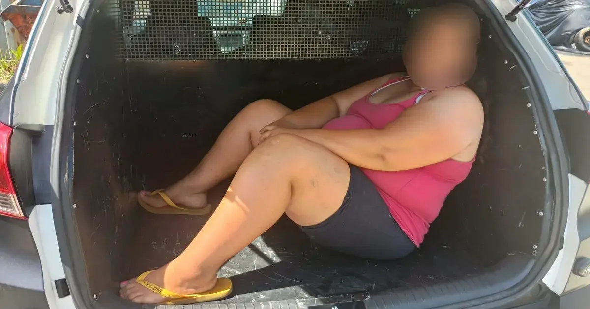 Pessoa sentada no porta-malas de um veículo, com os joelhos dobrados e vestindo camiseta e short.