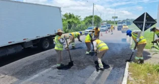 Pessoas realizando manutenção no asfalto da rodovia, com caminhões e cones de sinalização ao fundo.