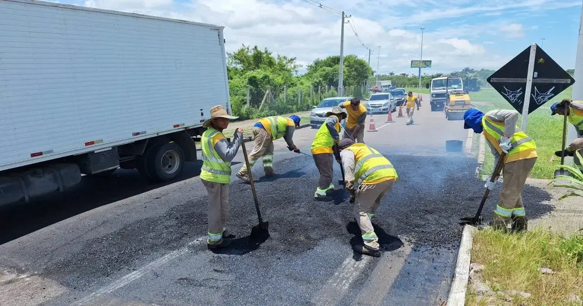 Pessoas realizando manutenção no asfalto da rodovia, com caminhões e cones de sinalização ao fundo.