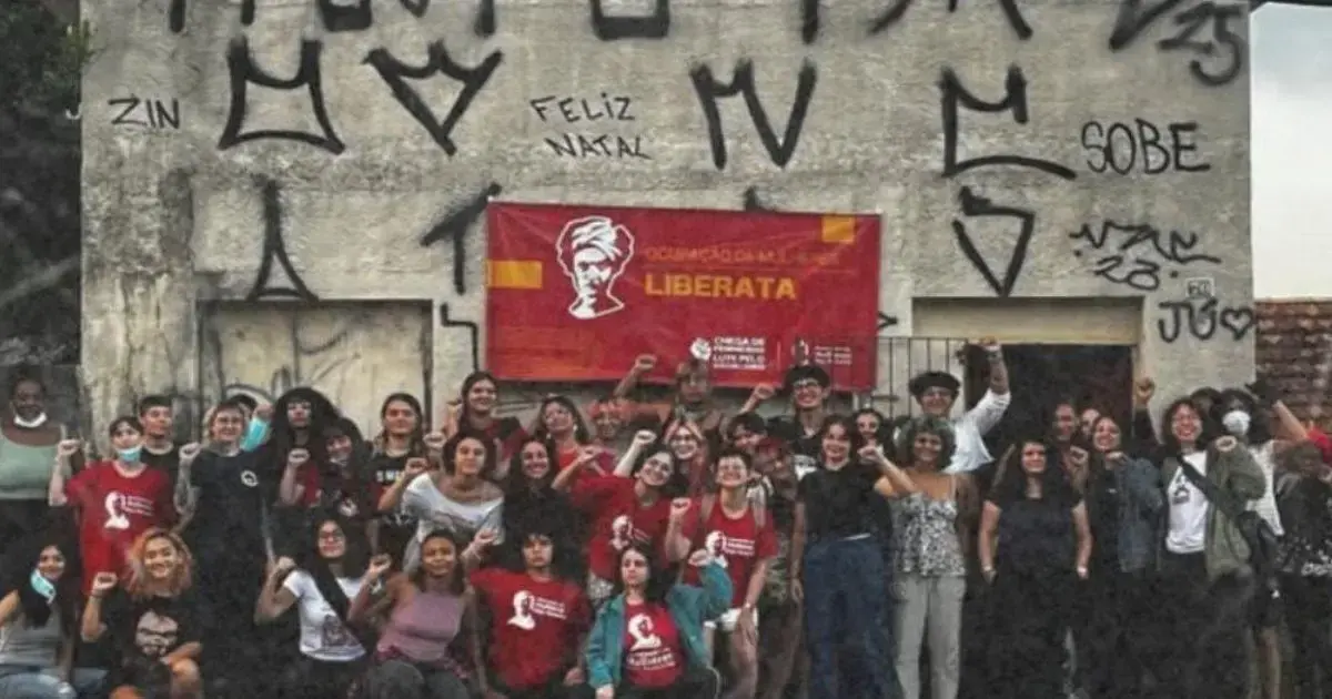 Grupo de pessoas em frente a um imóvel com grafites, segurando uma faixa com o texto 