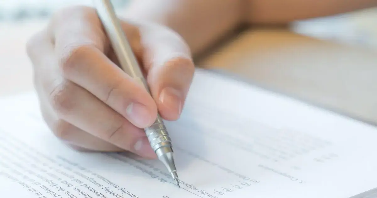 Mão segurando uma caneta prateada, escrevendo em uma folha com texto impresso.