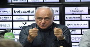 Homem falando em uma mesa com microfone, gesticulando, em frente a um painel com várias marcas.