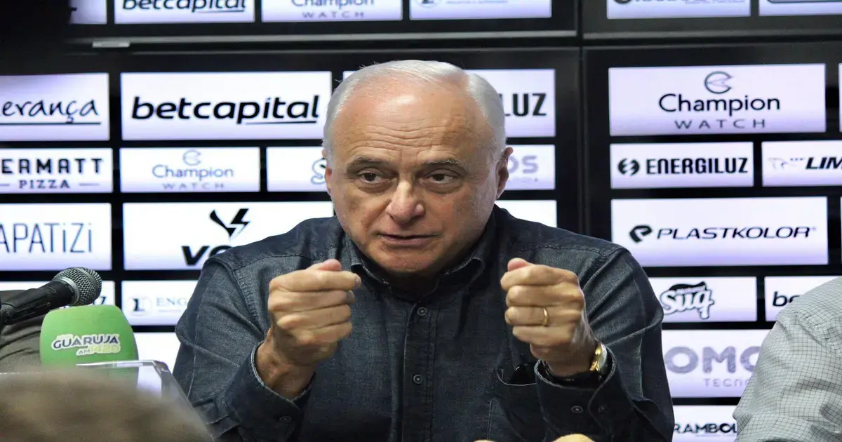 Homem falando em uma mesa com microfone, gesticulando, em frente a um painel com várias marcas.