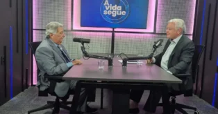 Dois homens sentados à mesa em um estúdio de gravação, conversando com microfones e painel de fundo iluminado.