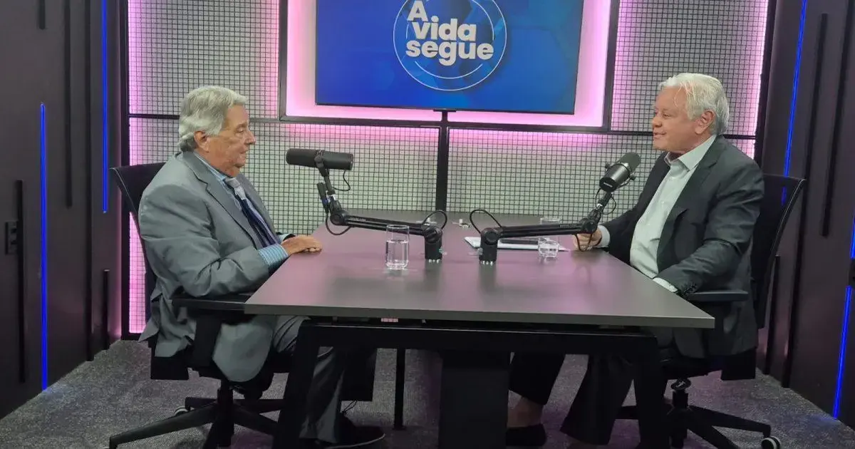 Dois homens sentados à mesa em um estúdio de gravação, conversando com microfones e painel de fundo iluminado.