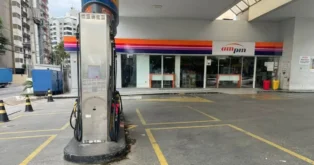 Posto de gasolina com bomba e loja de conveniência, apresentando carros estacionados e cones de sinalização.