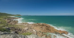 Vista de uma costa com rochas à beira-mar, mar azul e vegetação nas encostas.