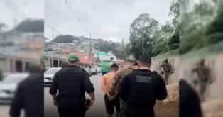 Grupo de policiais conduzindo um homem algemado em uma rua, com casas ao fundo.