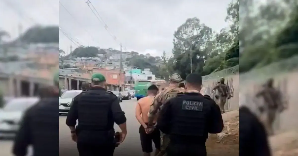 Grupo de policiais conduzindo um homem algemado em uma rua, com casas ao fundo.