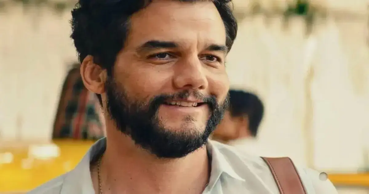 Sorriso de um homem com barba, vestindo uma camisa clara, em um fundo desfocado.