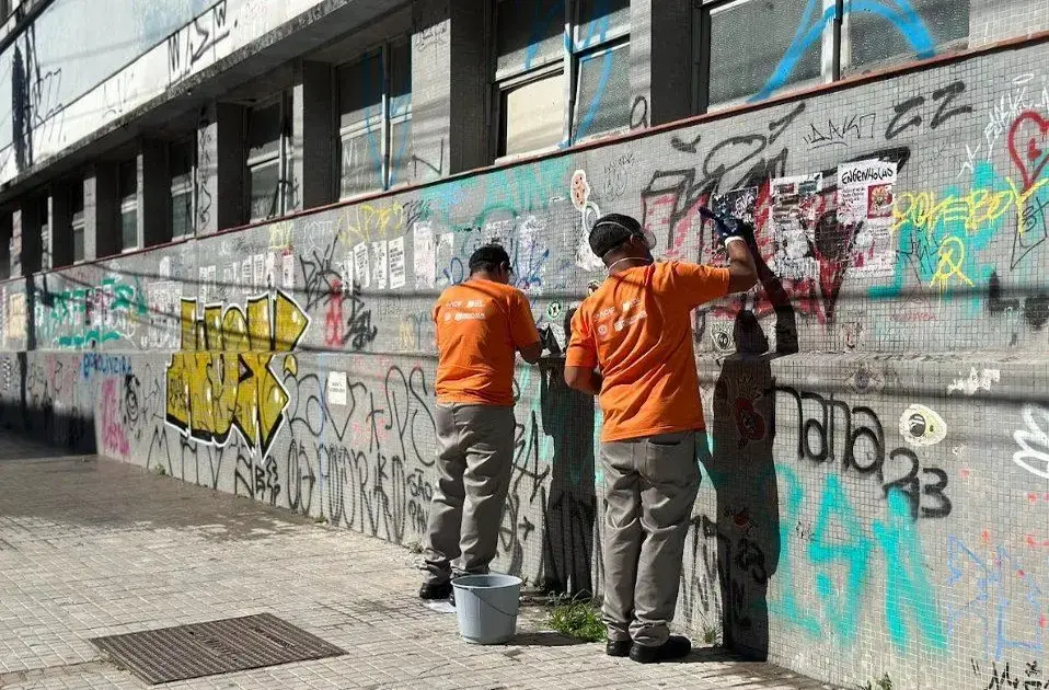Duas pessoas em uniformes laranja limpam graffiti de uma parede com tinta e adesivos visíveis.
