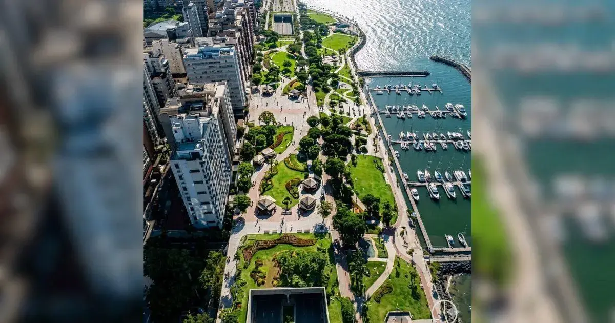 Vista aérea de um calçadão à beira-mar, com áreas verdes, calçadas e embarcações ancoradas.