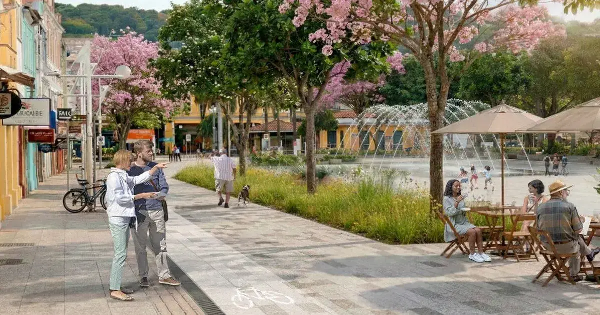 Caminho pavimentado em área com árvores, pessoas interagindo, pet, fonte e mesas ao ar livre.