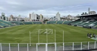 Gramado de um estádio de futebol, com arquibancadas ao fundo e várias edificações urbanas em volta.
