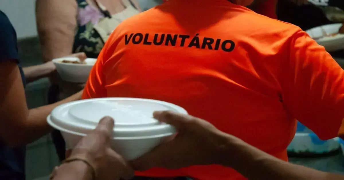 Voluntário em camisa laranja distribui marmitas em um ambiente interno. Pessoas ao fundo interagem.