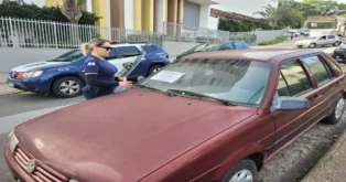 Mulher em uniforme de policiamento vistoria veículo da Volkswagem com aviso no para-brisa. Carro policial ao fundo.