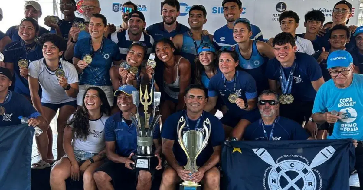 Grupo de atletas sorridentes exibindo medalhas e troféus, com bandeira do clube ao fundo.