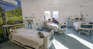 Quarto de hospital com leitos vazios e equipamentos médicos. Profissional de saúde organizando material ao lado de uma cama.