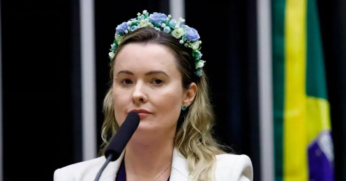 Mulher com cabelo solto e tiara de flores, falando ao microfone em um ambiente formal, com bandeira ao fundo.