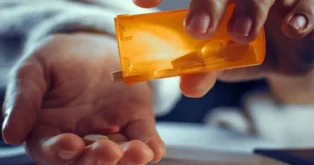 Mão segurando um frasco laranja de remédios, enquanto outra mão recebe várias pílulas.