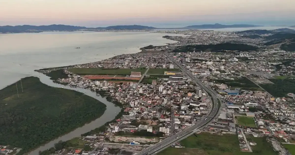 Vista aérea de uma cidade costeira, com áreas urbanas, estradas e vegetação ao redor.