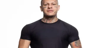 Indivíduo com tatuagens visíveis no rosto e no pescoço, vestindo camiseta preta.