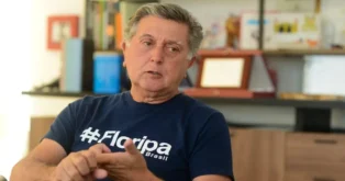 Homem conversando, vestindo camiseta azul com a inscrição "#Floripa Brasil", em ambiente interno com prateleiras ao fundo.