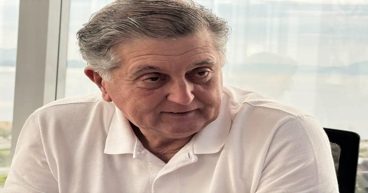 Homem com cabelo grisalho e roupa branca, olhando para a câmera com expressão concentrada em ambiente interno.