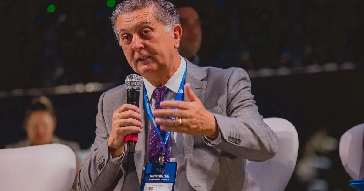 Homem com terno cinza falando em um evento, segurando um microfone e gesticulando com a mão.