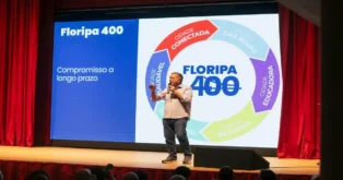 Pessoa em pé apresentando um projeto chamado "Floripa 400", com texto sobre compromisso a longo prazo e elementos visuais de várias cidades.