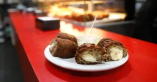 Prato com almôndegas ao meio, mostrando recheio cremoso, acompanhado de um recipiente com molho em mesa vermelha.