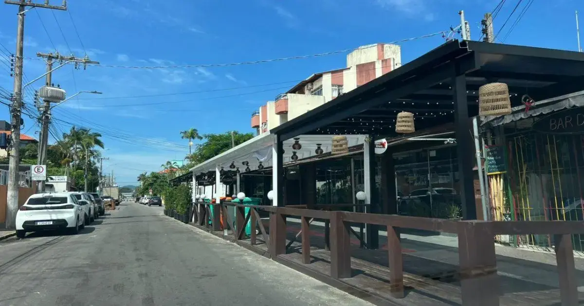 Rua com restaurante à direita, mesas externas, árvores e céu azul ao fundo. Veículos estacionados na calçada.