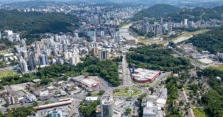 Vista aérea de uma cidade com prédios, áreas verdes, um rio e uma rodovia visível.