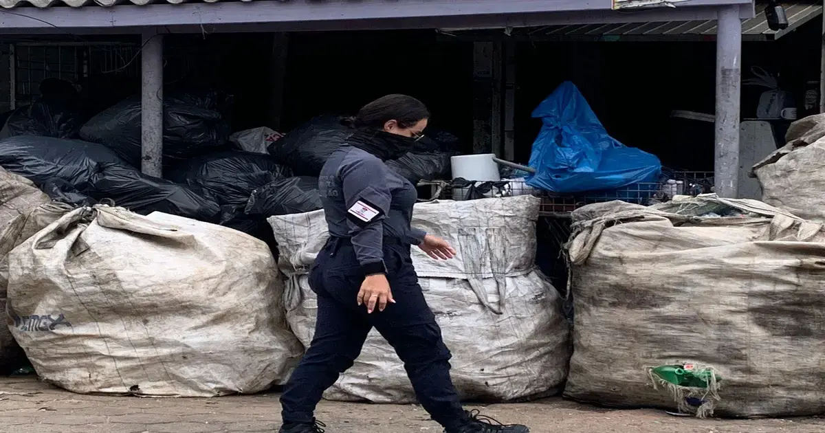 Pessoa usando uniformes escuros, caminhando ao lado de sacos de lixo grandes e outros objetos acumulados.
