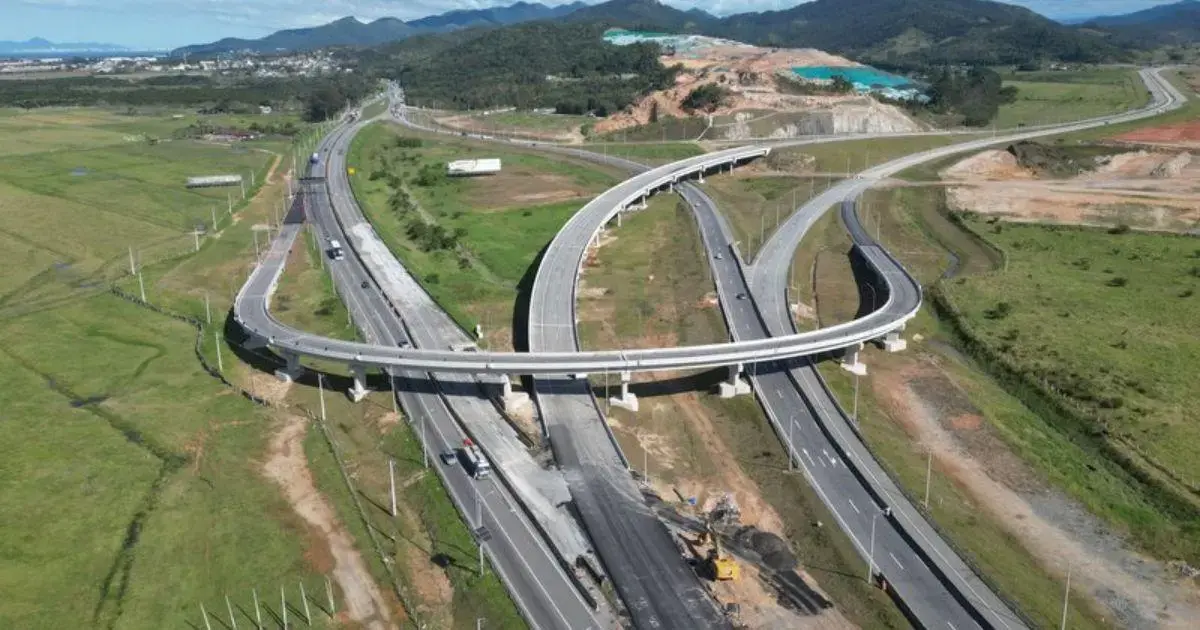 Vista aérea de uma interseção de rodovias com obras em andamento e áreas verdes ao fundo.