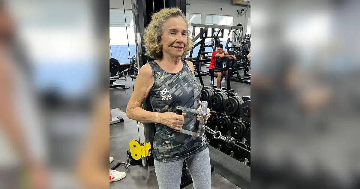 Mulher de cabelo cacheado em academia segurando equipamento de musculação, com pesos ao fundo.